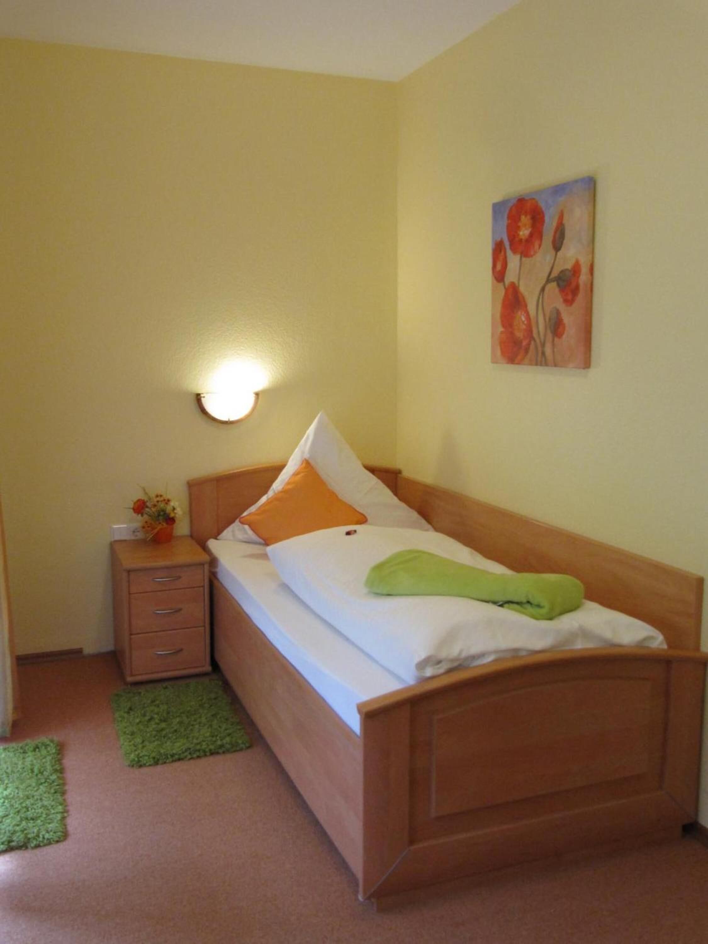 A small, neatly arranged bedroom with a single bed, a nightstand, a green rug, and a wall-mounted light.