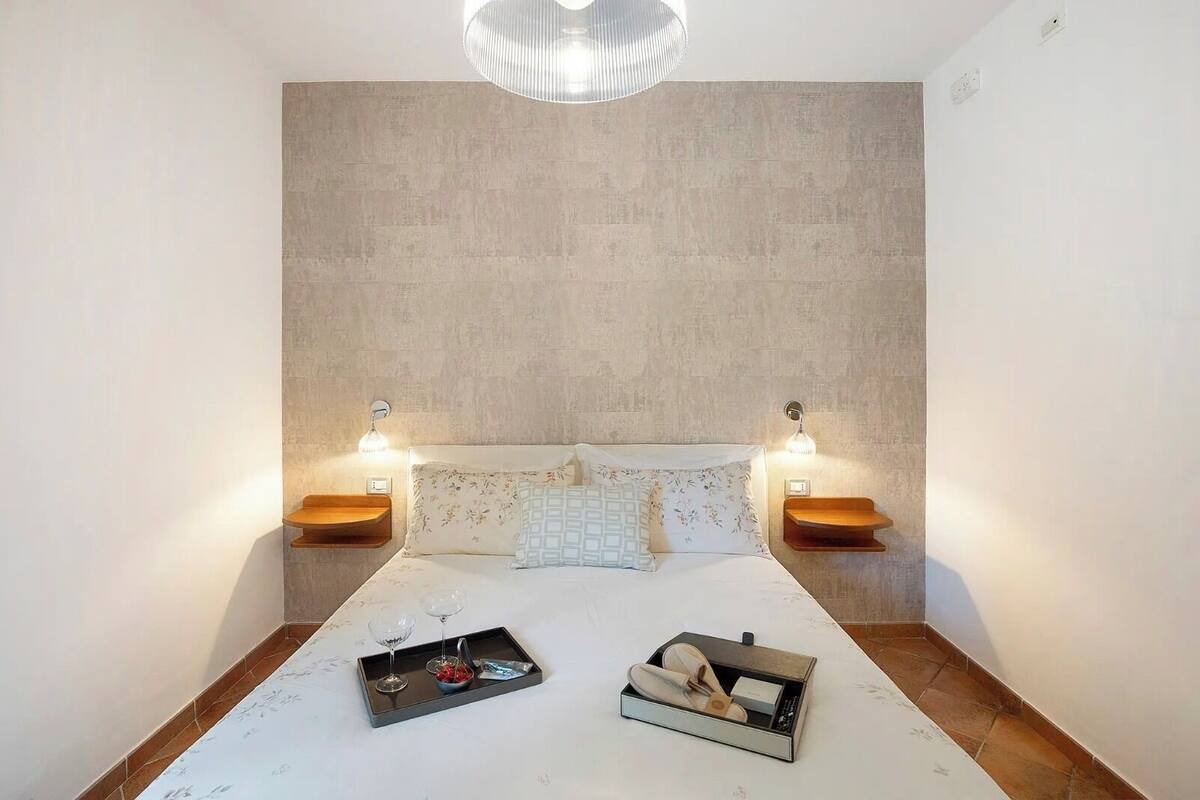 Een slaapkamer met een groot bed, twee nachtkastjes, een hanglamp en een dienblad met wijnglazen en een doos op het bed.