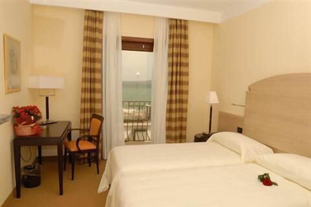 Habitación de hotel con cama, escritorio, silla, lámpara y vista al mar por la ventana.