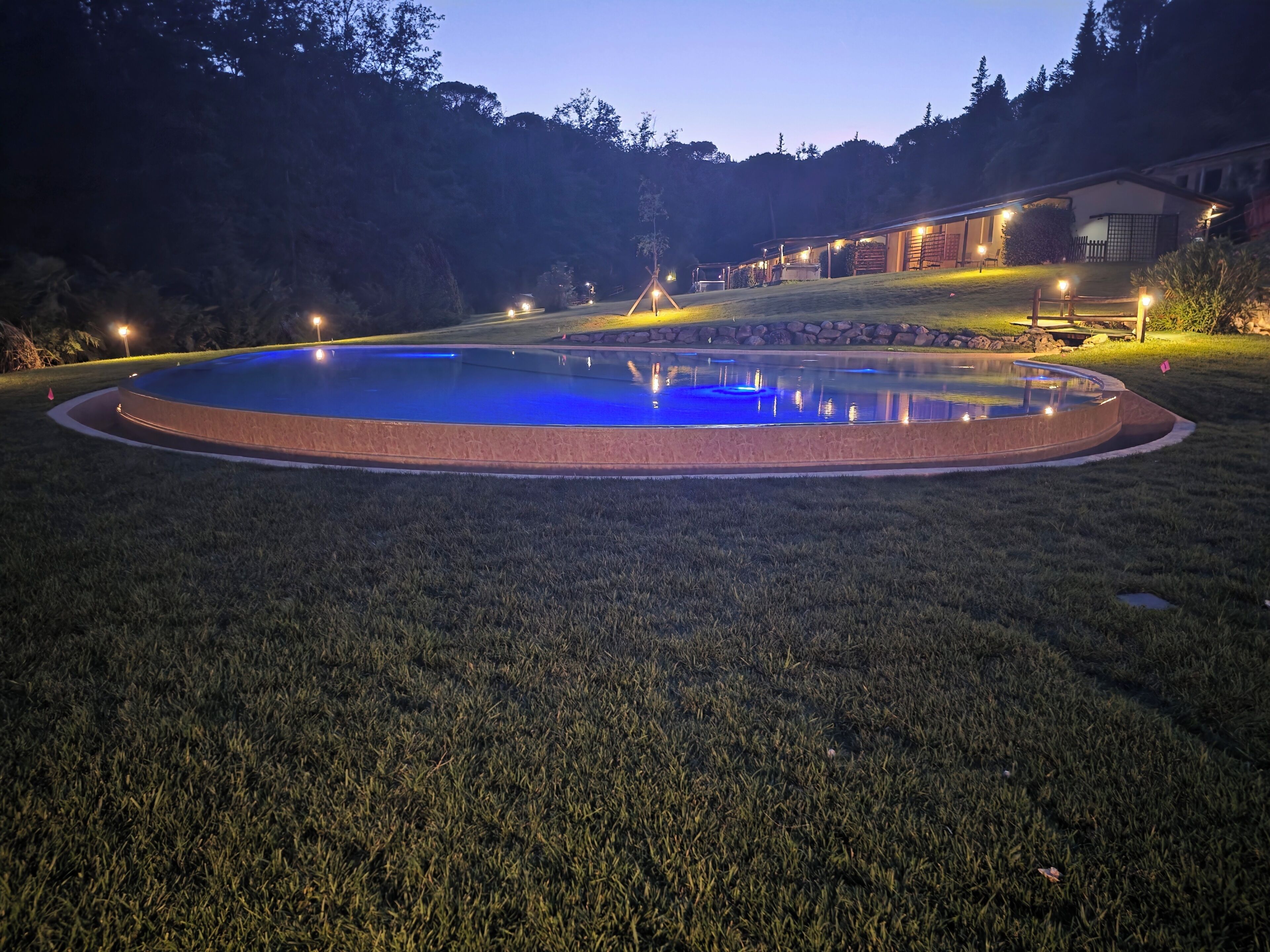 A circular swimming pool with a surrounding stone edge, set in a grassy area with a building in the background.