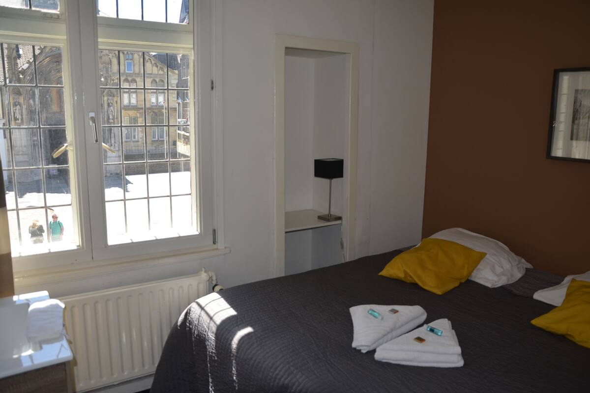 Habitación de hotel con un ventanal grande, una cama con almohadas amarillas, una mesita de noche con una lámpara y un cuadro enmarcado en la pared.