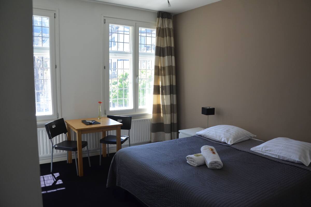 Habitación de hotel con cama, escritorio y silla. Cuenta con ventana con cortinas, radiador y lámpara de pared.