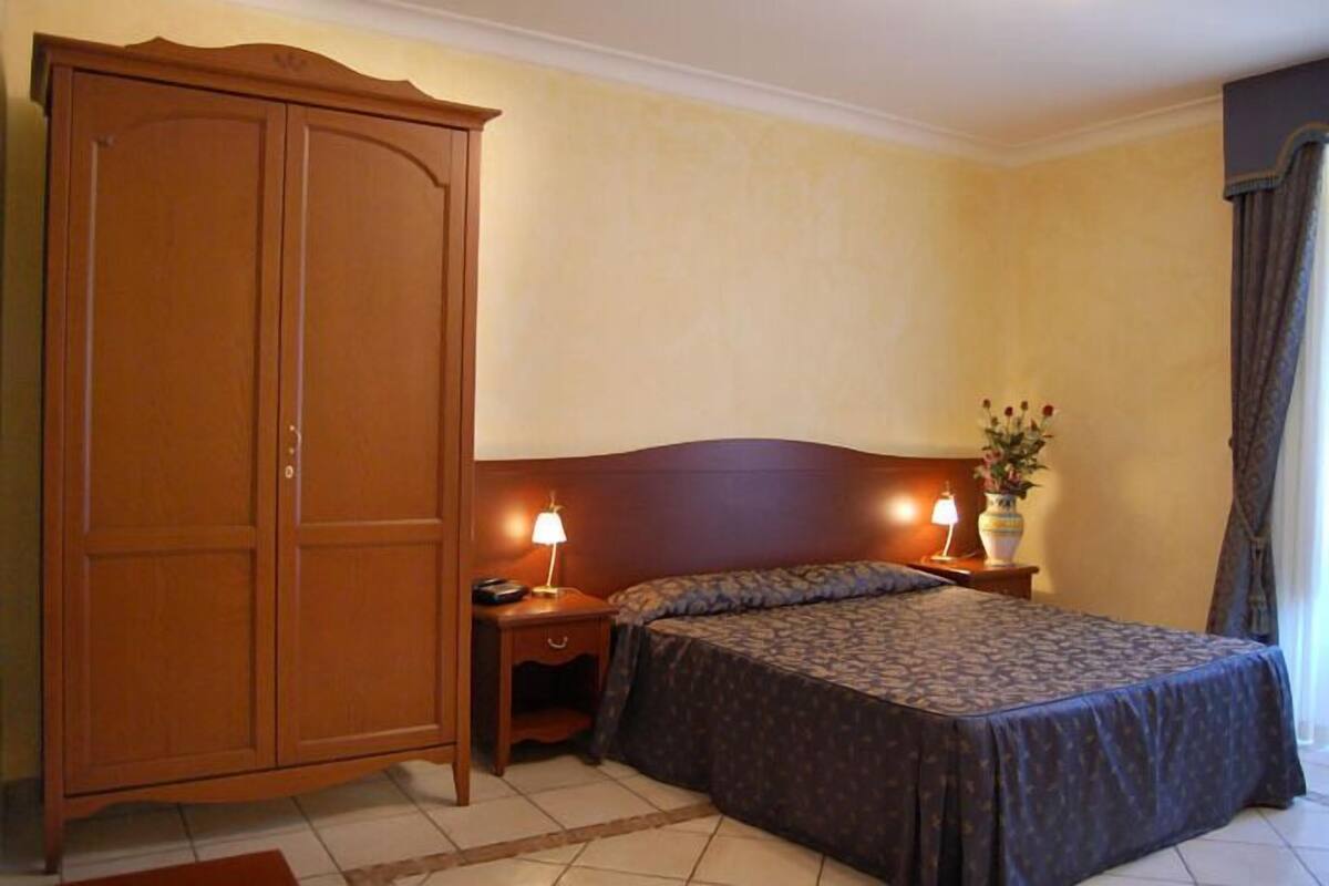 A bedroom with a double bed, a nightstand with a lamp, a wooden wardrobe, and a vase with flowers on the nightstand.