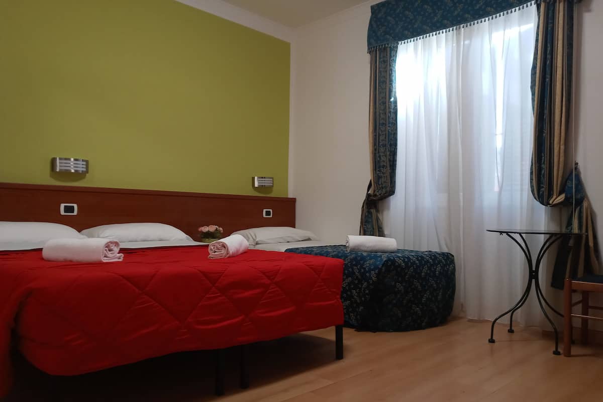 A hotel room with two beds, one with a red bedspread and the other with a blue patterned bedspread. There is a small table with a chair, a window with sheer curtains, and a wall-mounted air conditioning unit.