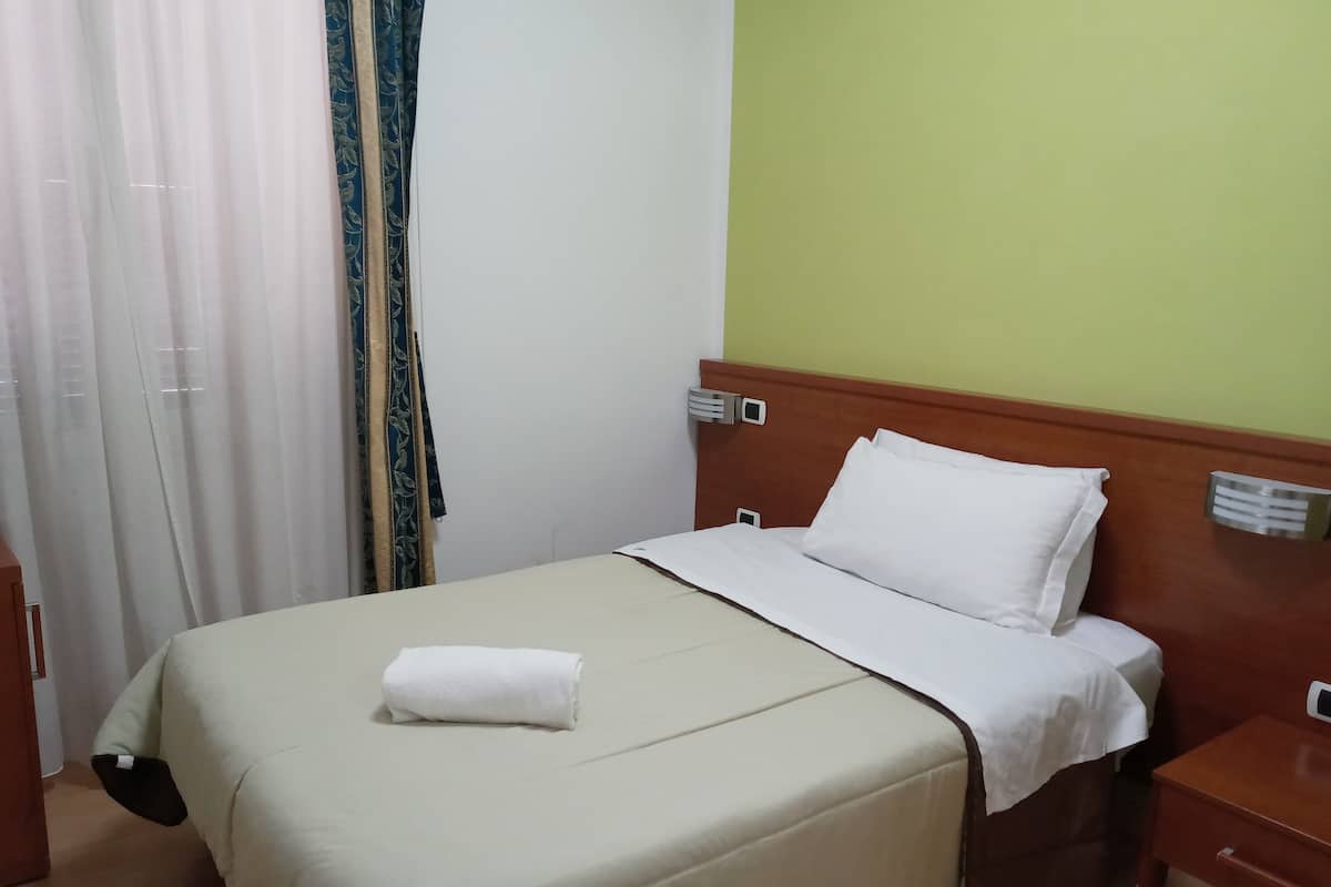 A neatly made bed with a white comforter and pillows, a wooden nightstand with a lamp, and a window with curtains.