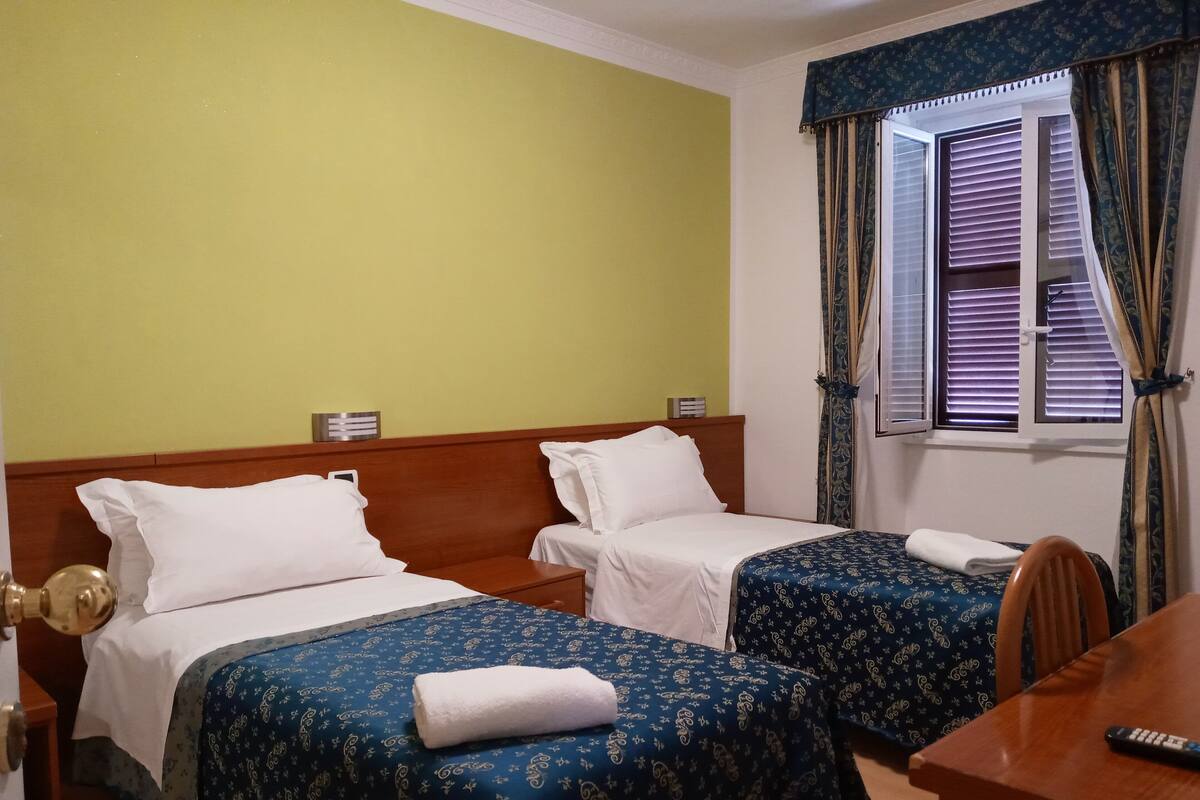 A hotel room with two single beds, a wooden headboard, a desk with a remote control, and a window with curtains.