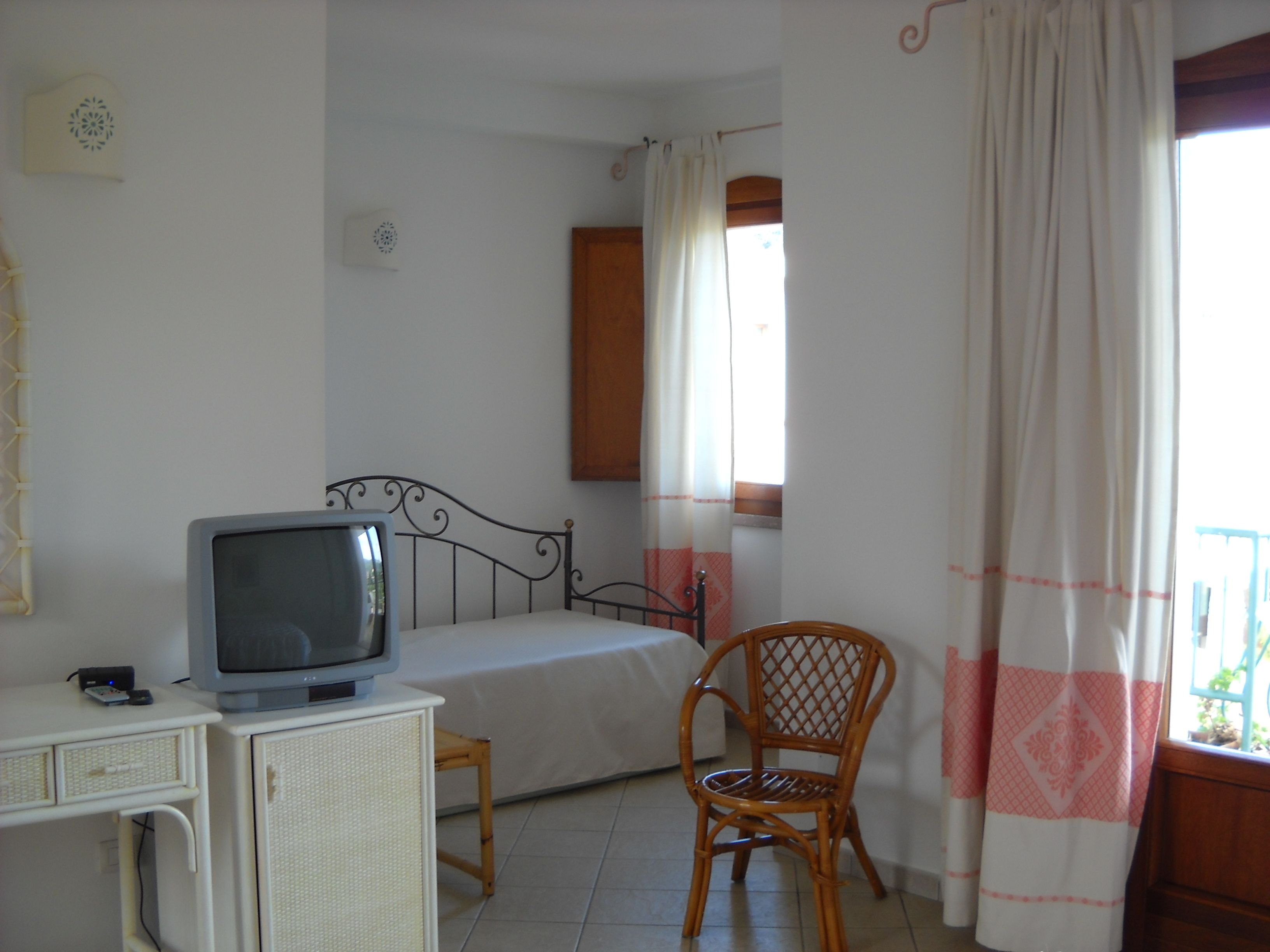 Quarto com cama, televisão sobre suporte, cadeira e janela com cortinas.