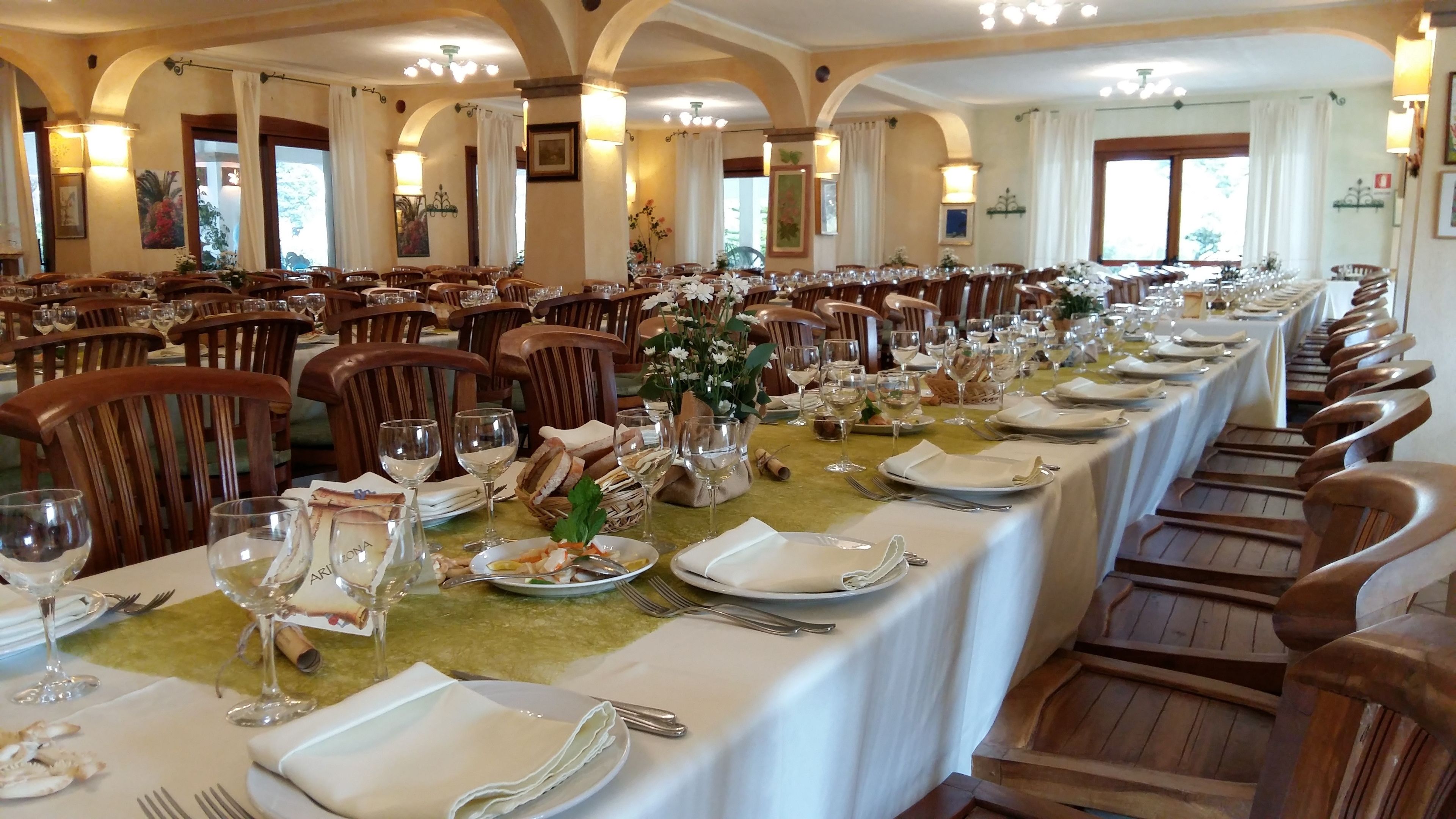 A long dining table set with white tablecloths, wine glasses, plates, and wooden chairs in a well-lit room with arched ceilings and framed artwork on the walls.