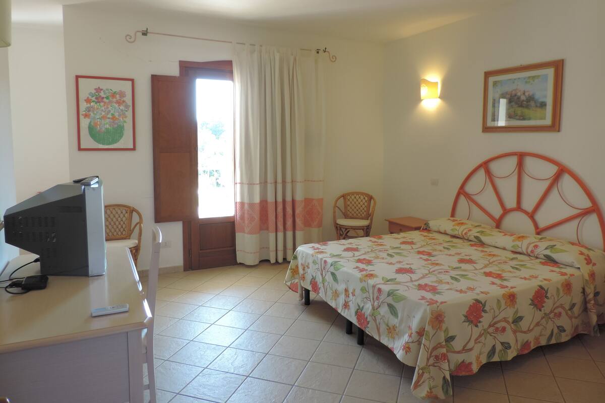 Ein Schlafzimmer mit Bett, Schreibtisch mit Computer, Stuhl, Fenster mit Vorhängen und einer Wandleuchte.