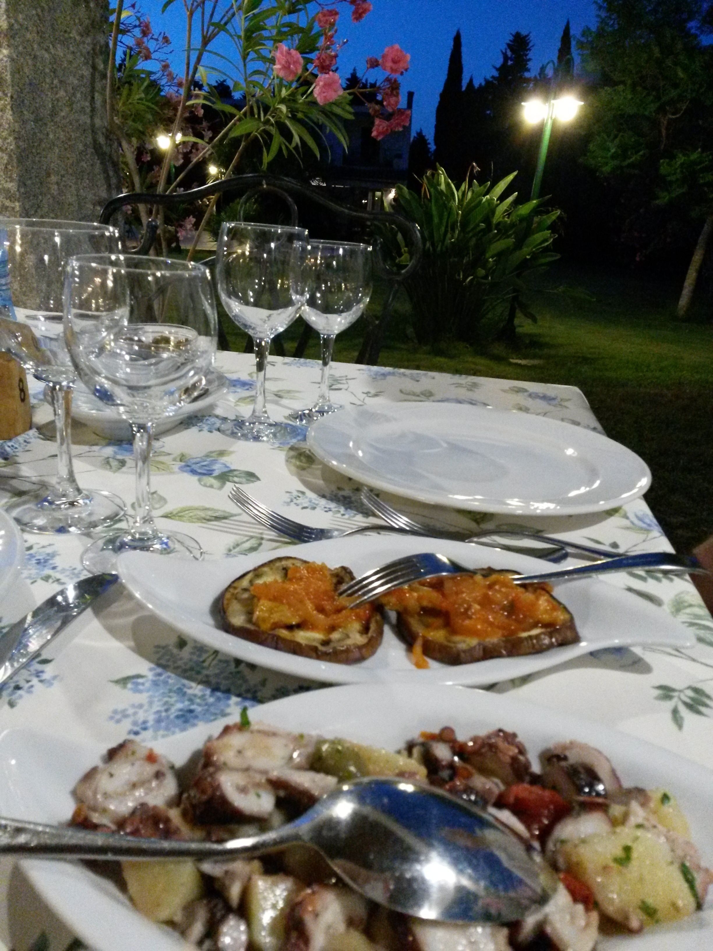 A table set outdoors with plates of food, wine glasses, and cutlery.