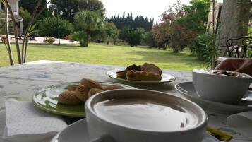Una mesa con una taza de café, un plato de galletas y un tazón de sopa, colocada al aire libre con vista a un jardín y árboles.