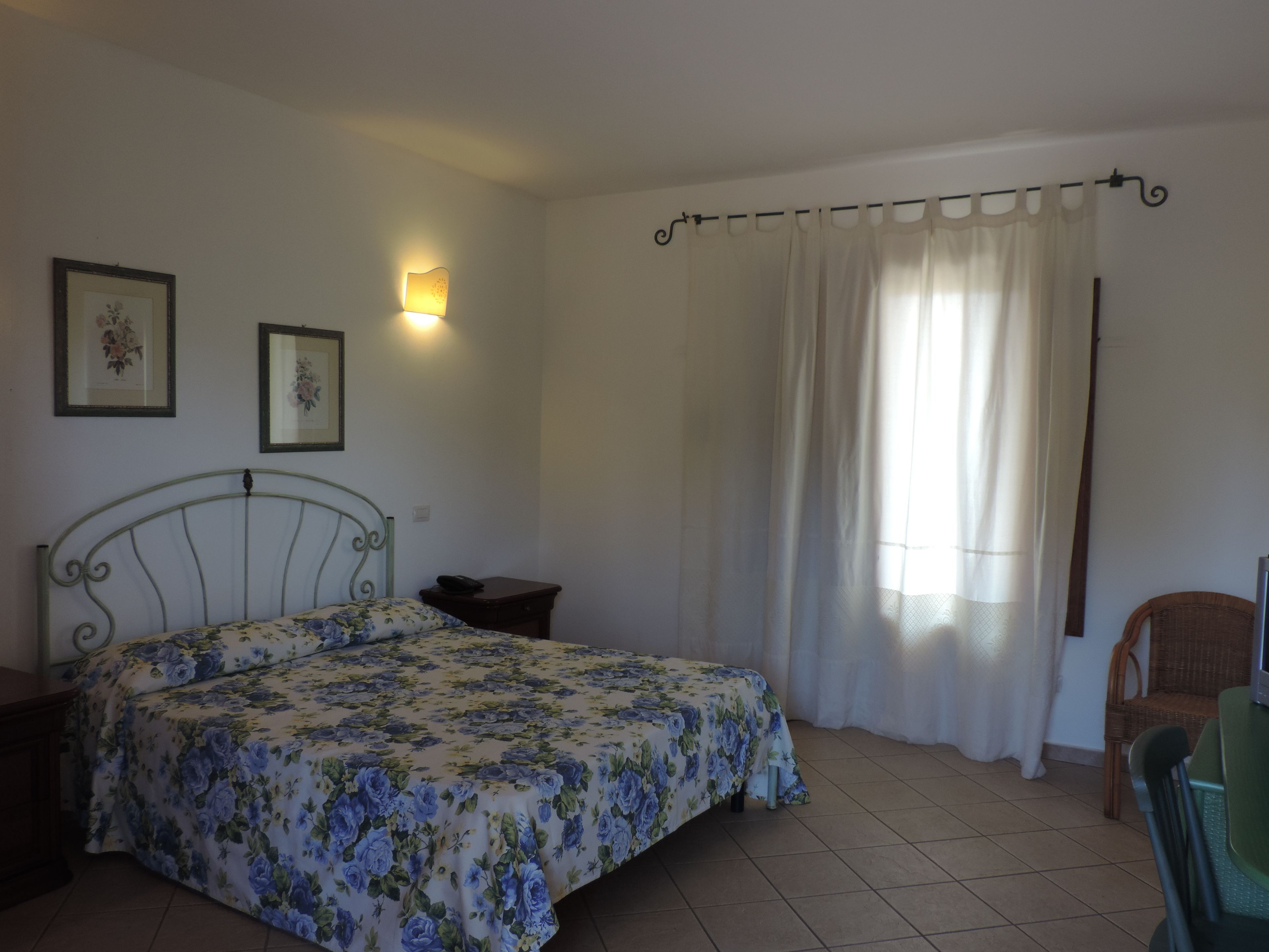 Un dormitorio con cama, una silla, una mesita de noche, una lámpara de pared y una ventana con cortinas.