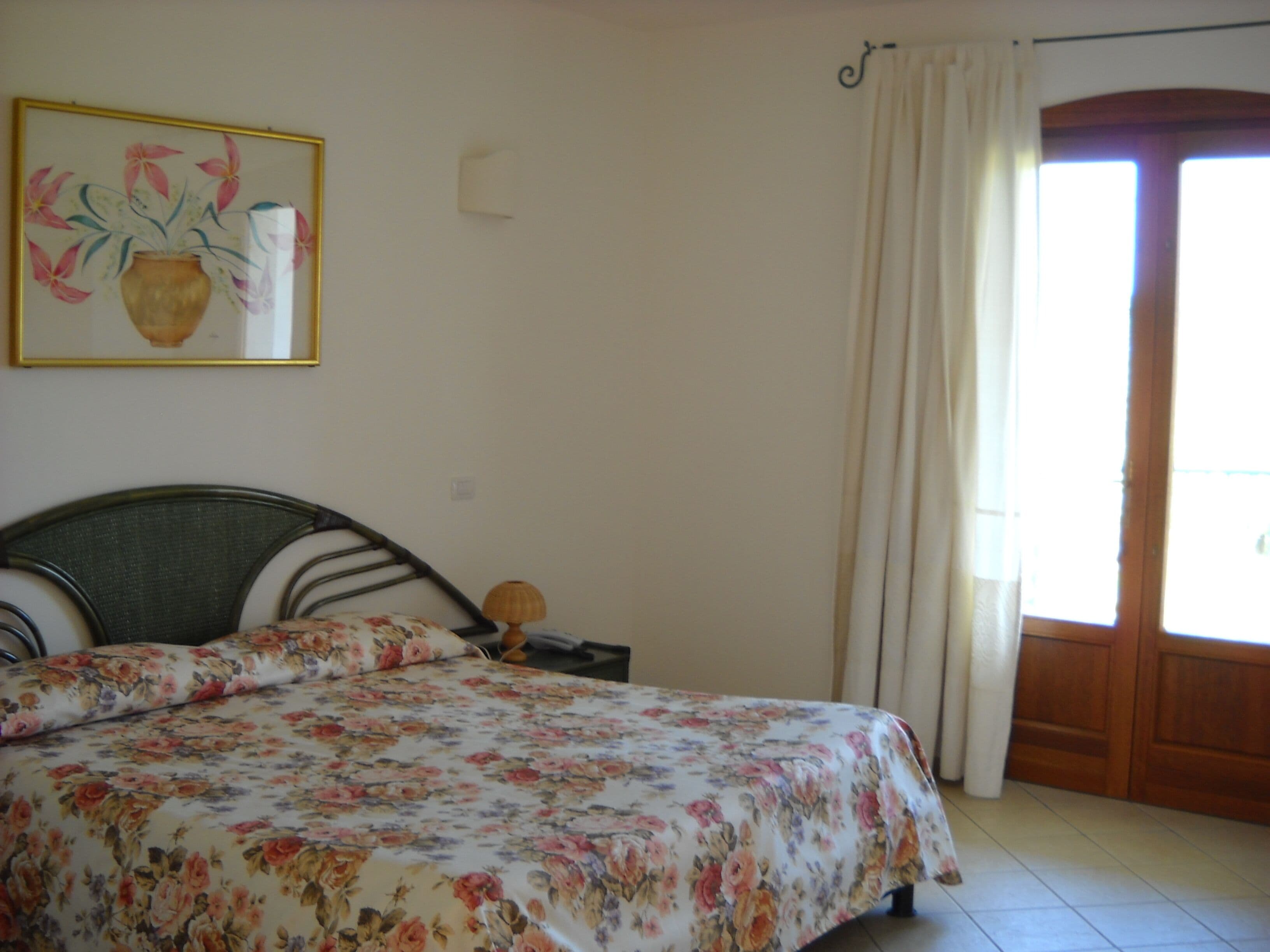 A bedroom with a floral bedspread, a headboard, a bedside table with a lamp, and a door with curtains.