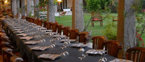 A long dining table set for a meal with chairs around it, under a covered area with columns and plants.