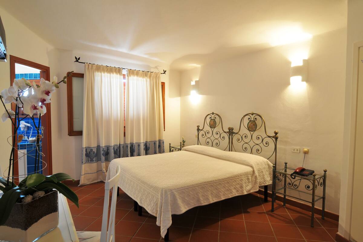 A bedroom with a bed, curtains, a wall-mounted light fixture, and a small table with a telephone.
