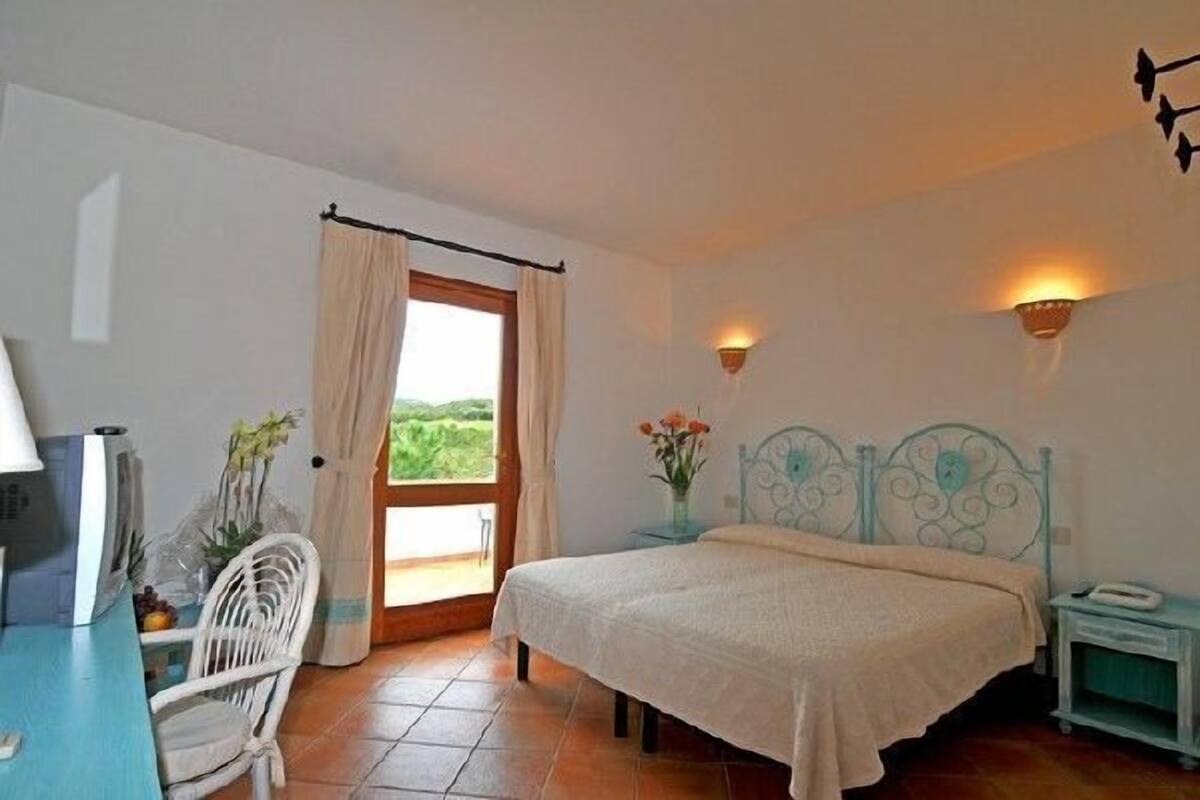 A bedroom with a bed, a desk, a chair, a window with curtains, and two wall-mounted lamps.