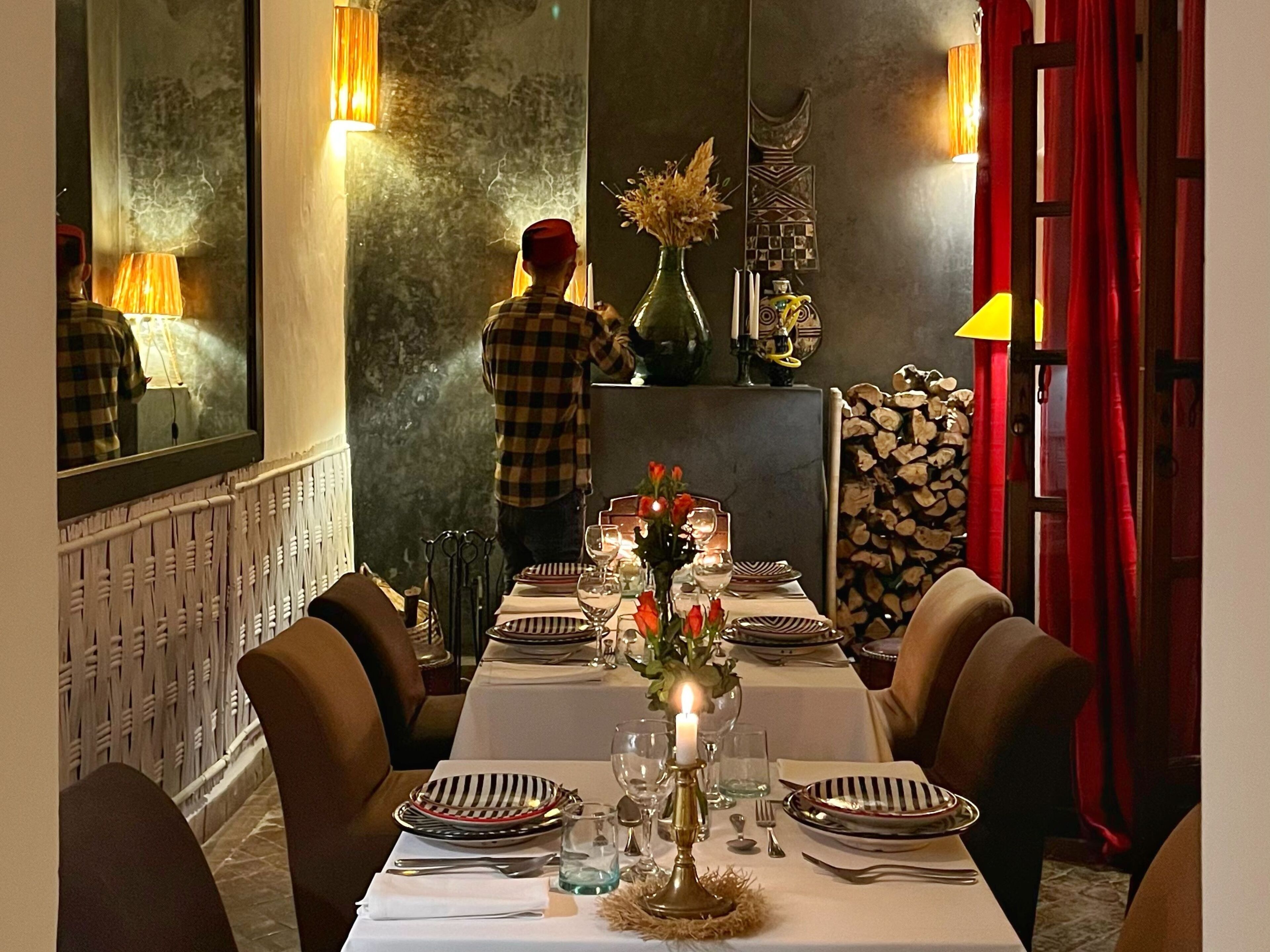 Un coin repas avec une longue table dressée, un miroir accroché au mur et une personne debout à l’arrière-plan.