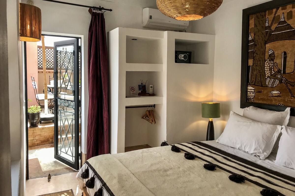 Une chambre à coucher comprenant un lit, une lampe suspendue, un climatiseur fixé au mur et une porte donnant sur un espace extérieur avec une table et des chaises.