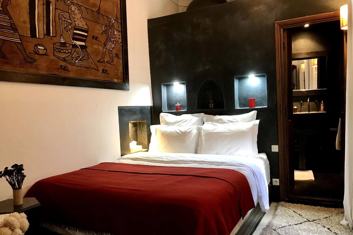 Une chambre d’hôtel avec un grand lit, une couverture rouge, une tête de lit fixée au mur, des lampes de chevet et un tableau décoratif.
