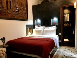 A hotel room with a large bed, a red blanket, a wall-mounted headboard, bedside lamps, and a decorative painting.