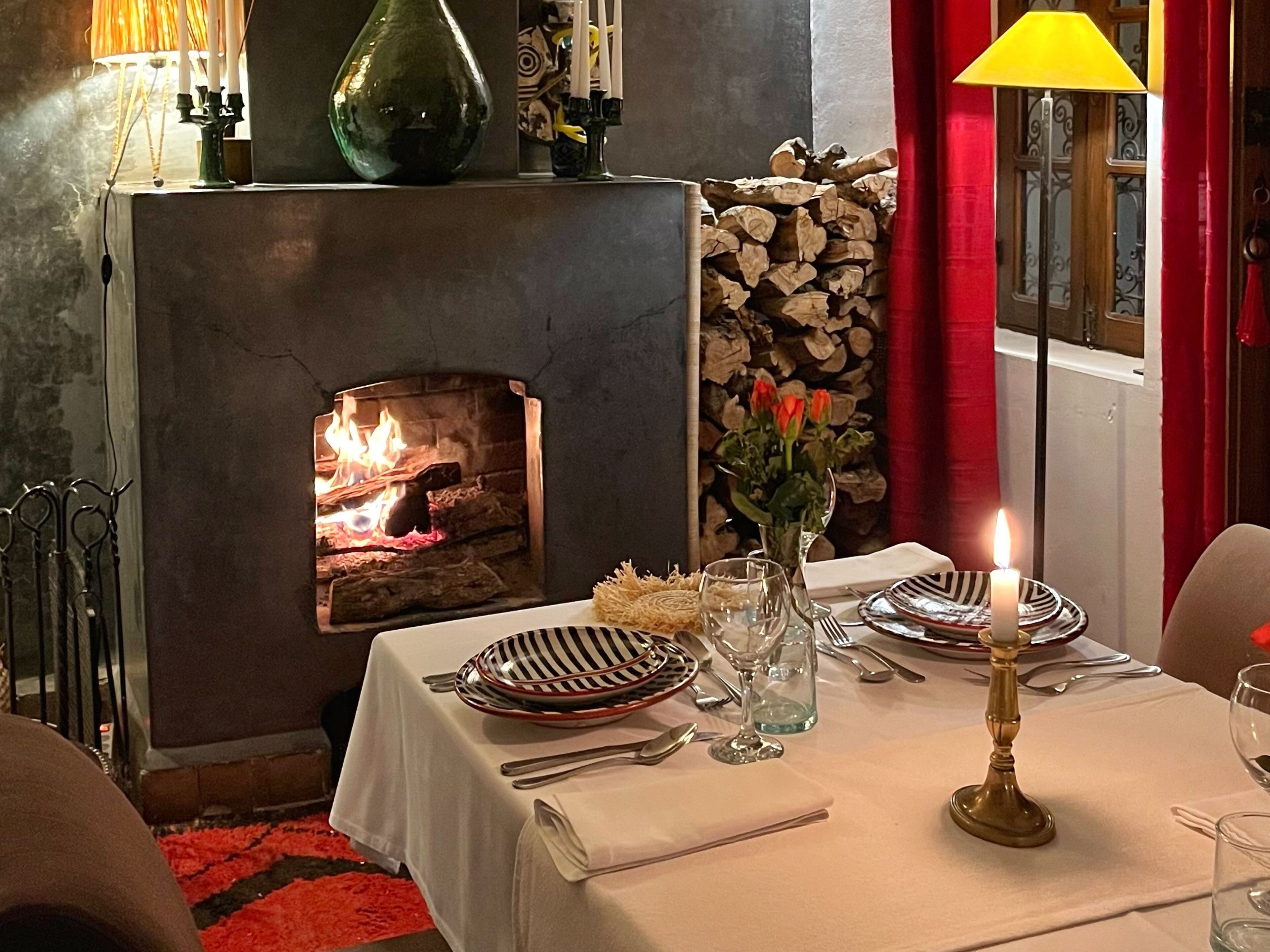 Un coin repas agrémenté d’une cheminée, d’une table dressée pour deux personnes et d’un vase rempli de fleurs.