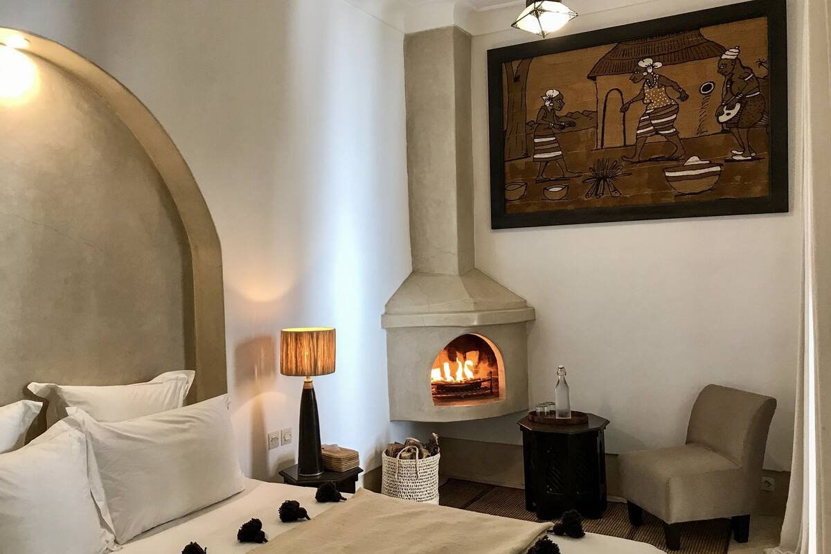 Une chambre avec une cheminée en pierre, un lit recouvert de draps blancs, une table de chevet avec une lampe et une œuvre d’art encadrée accrochée au mur.