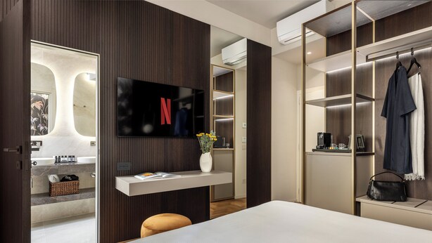 Ein modernes Hotelzimmer mit einem großen Bett, einem an der Wand befestigten Fernseher, einem Schminktisch mit Spiegel und einem begehbaren Kleiderschrank mit Kleidung.