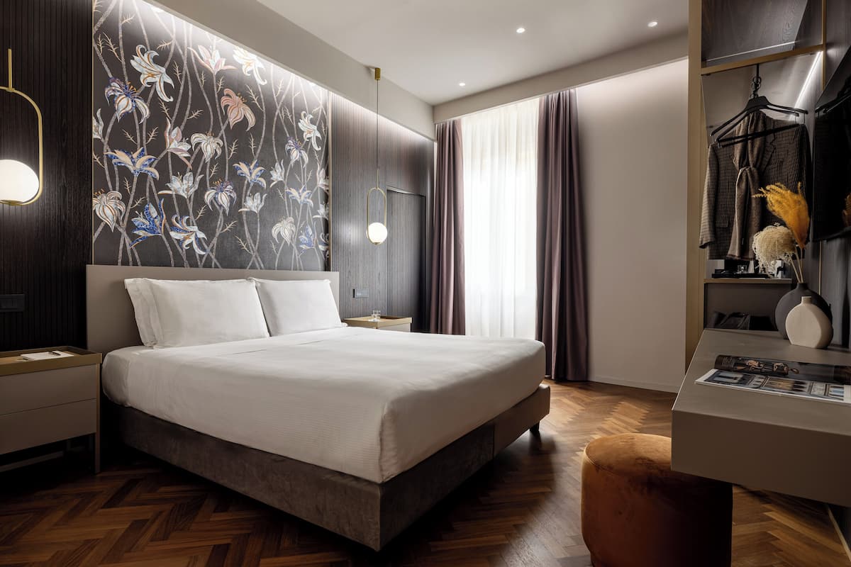 A modern hotel room with a large bed, a wooden floor, a wardrobe with hanging clothes, a desk with a lamp, and a wall with a floral wallpaper.