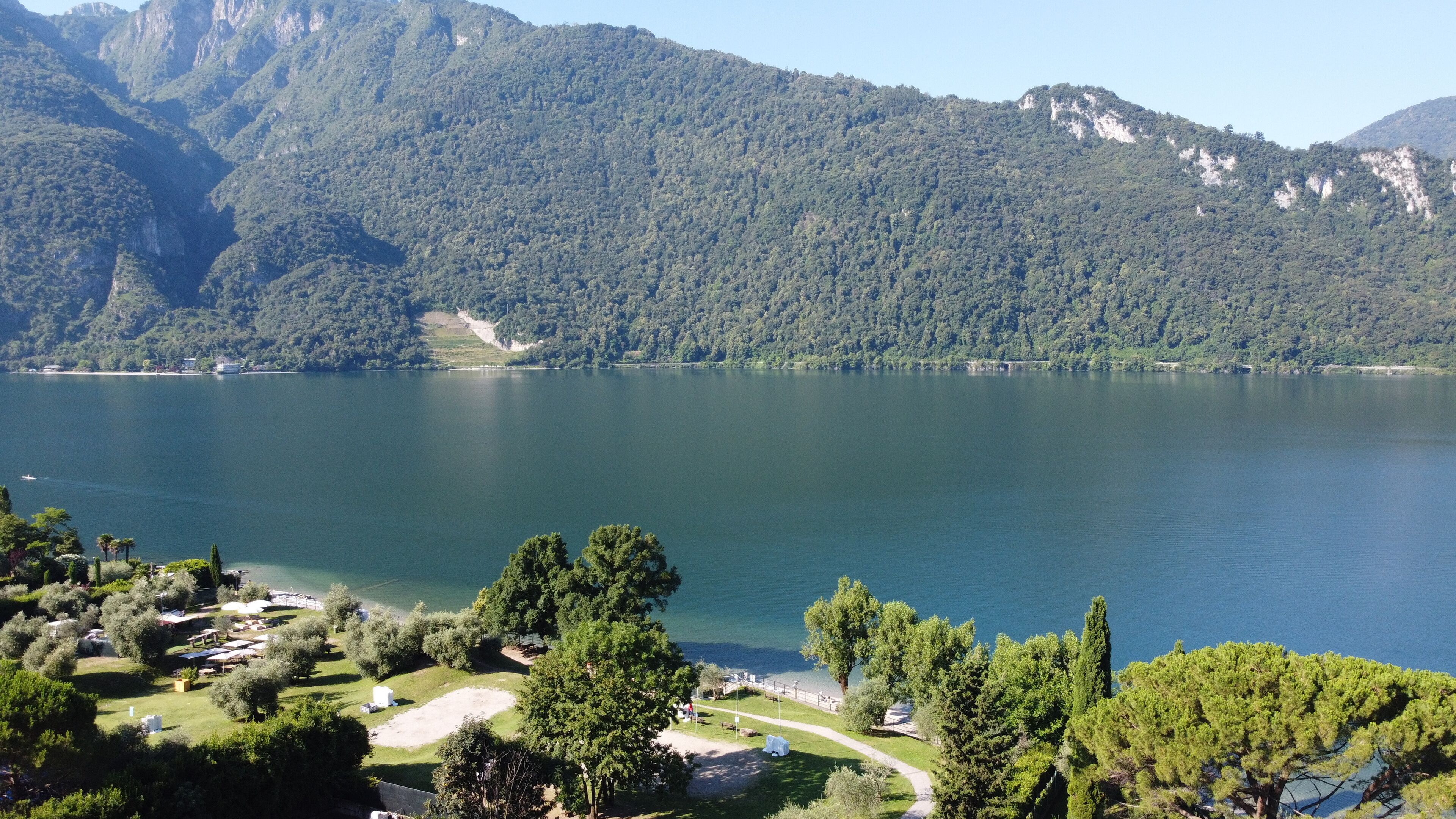 A lakeside view with a mountainous backdrop, a clear sky, and a small park area with trees and a pathway.
