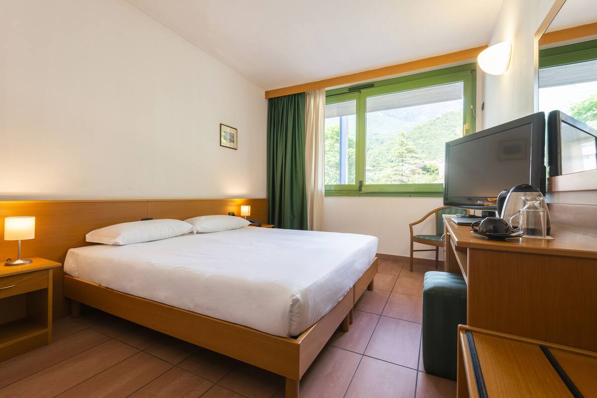 Habitación de hotel con cama, escritorio con televisor, una silla y ventana con cortinas.