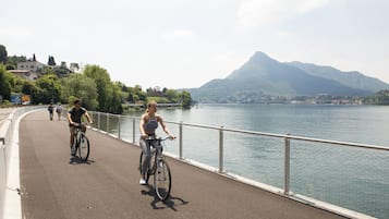 Une piste cyclable asphaltée longe un plan d’eau, avec des montagnes en arrière-plan.