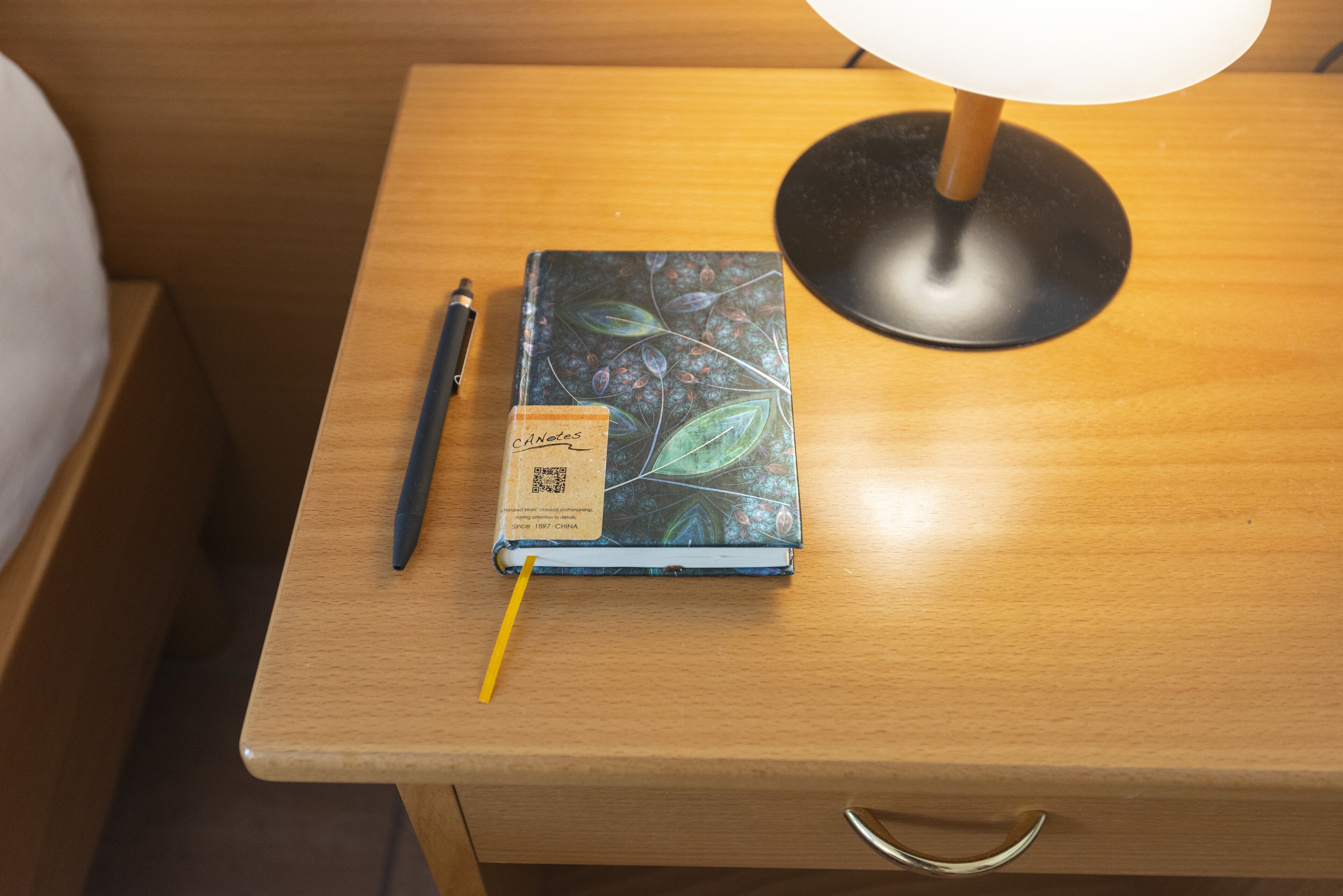 Een houten nachtkastje met een notitieboekje, pen en een lamp.