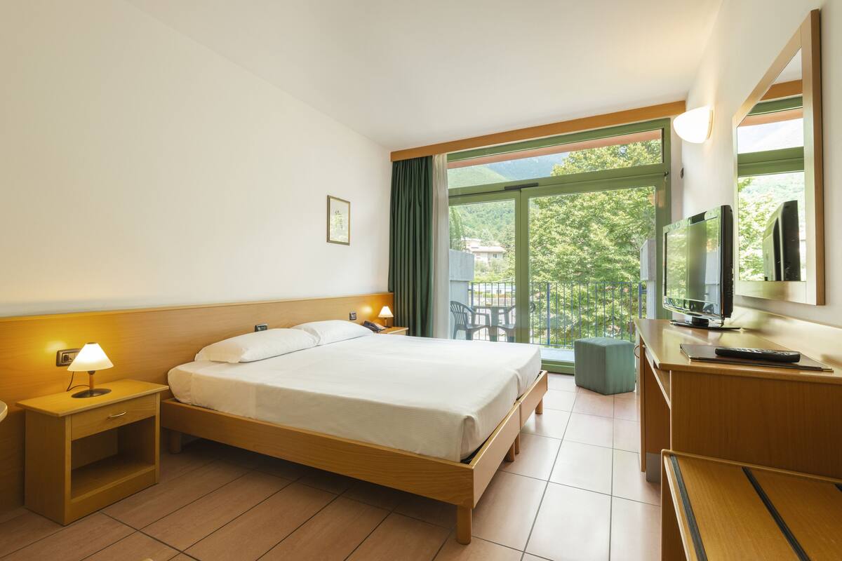 Habitación de hotel con cama, mesita de noche con lámpara, televisión y balcón con sillas.