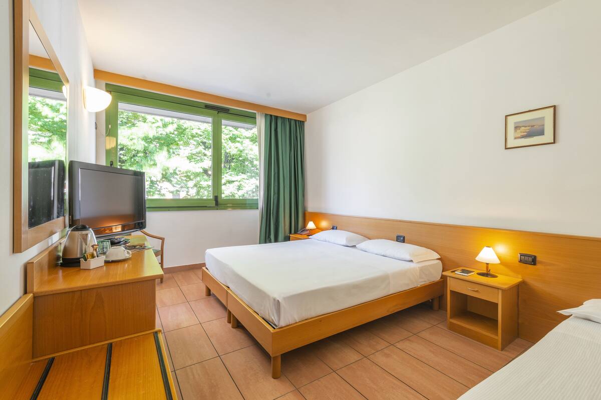 Una habitación de hotel con cama, mesitas de noche, televisión y ventana con cortinas.