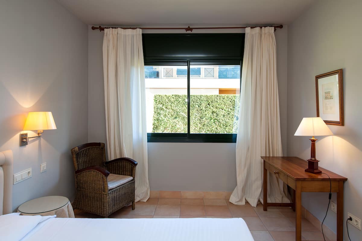 Een hotelkamer met een bed, een stoel, een bureau, twee lampen en een raam met gordijnen.