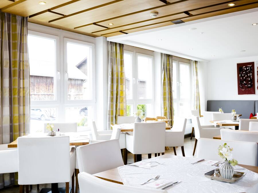 Uma sala de jantar com cadeiras e mesas brancas, janelas amplas com cortinas e um design de teto moderno.