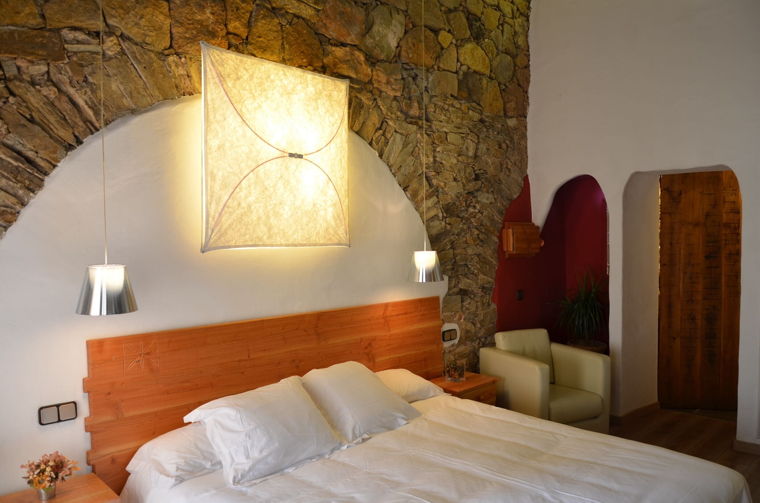 A bedroom with a stone wall, a wooden headboard, a bed with white linens, a nightstand, a lamp, and a chair.