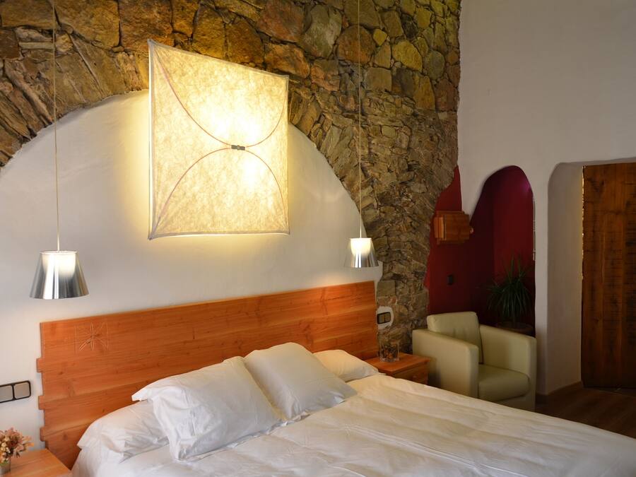 Un dormitorio con pared de piedra, cabecera de madera, cama con ropa blanca, mesita de noche, lámpara y una silla.