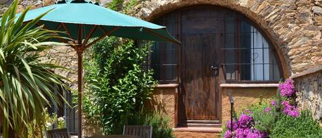 A stone building with a wooden door and a green umbrella. There are chairs and a table outside.