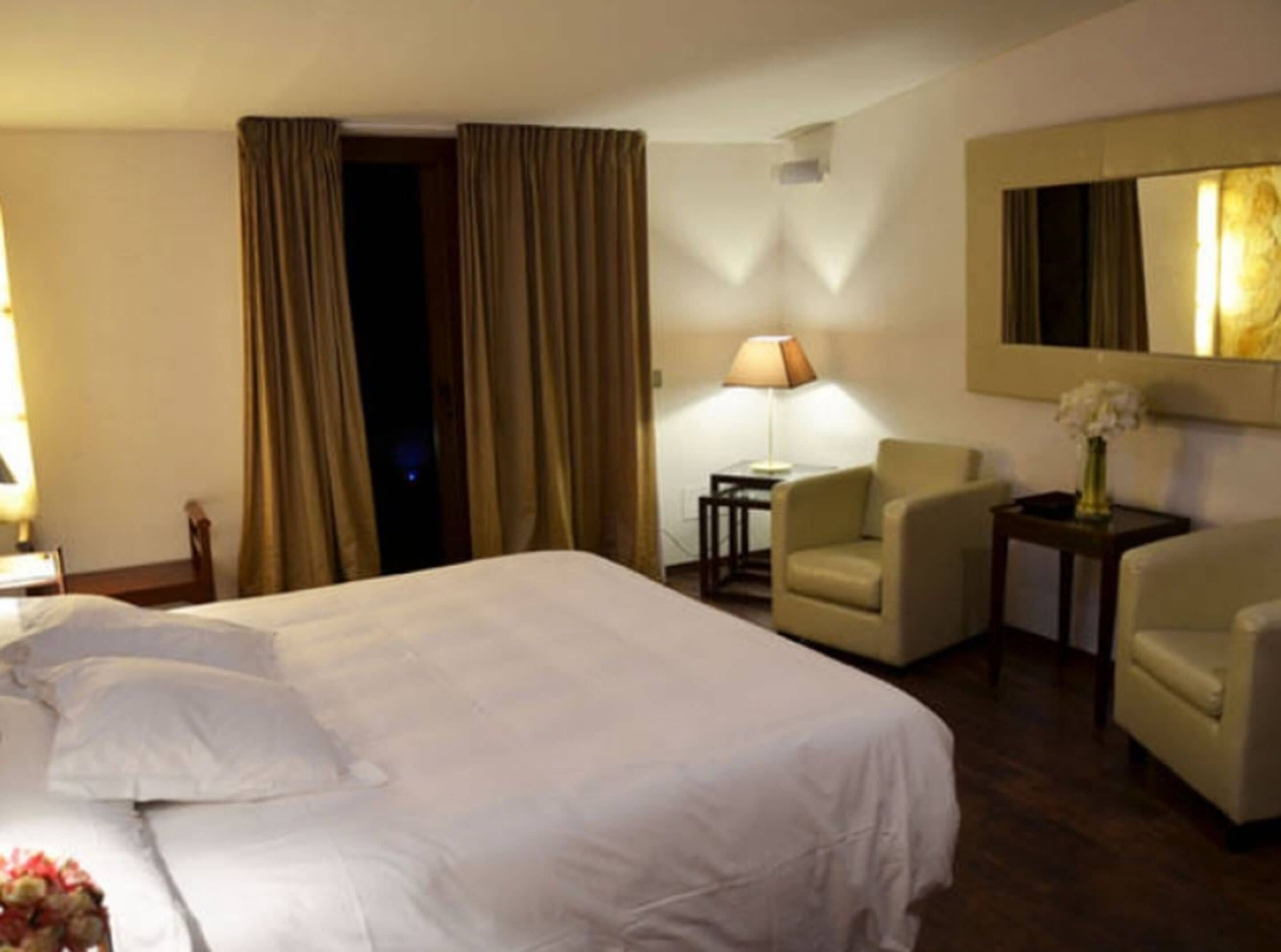 A hotel room with a large bed, two armchairs, a small table with a lamp, and a mirror on the wall.