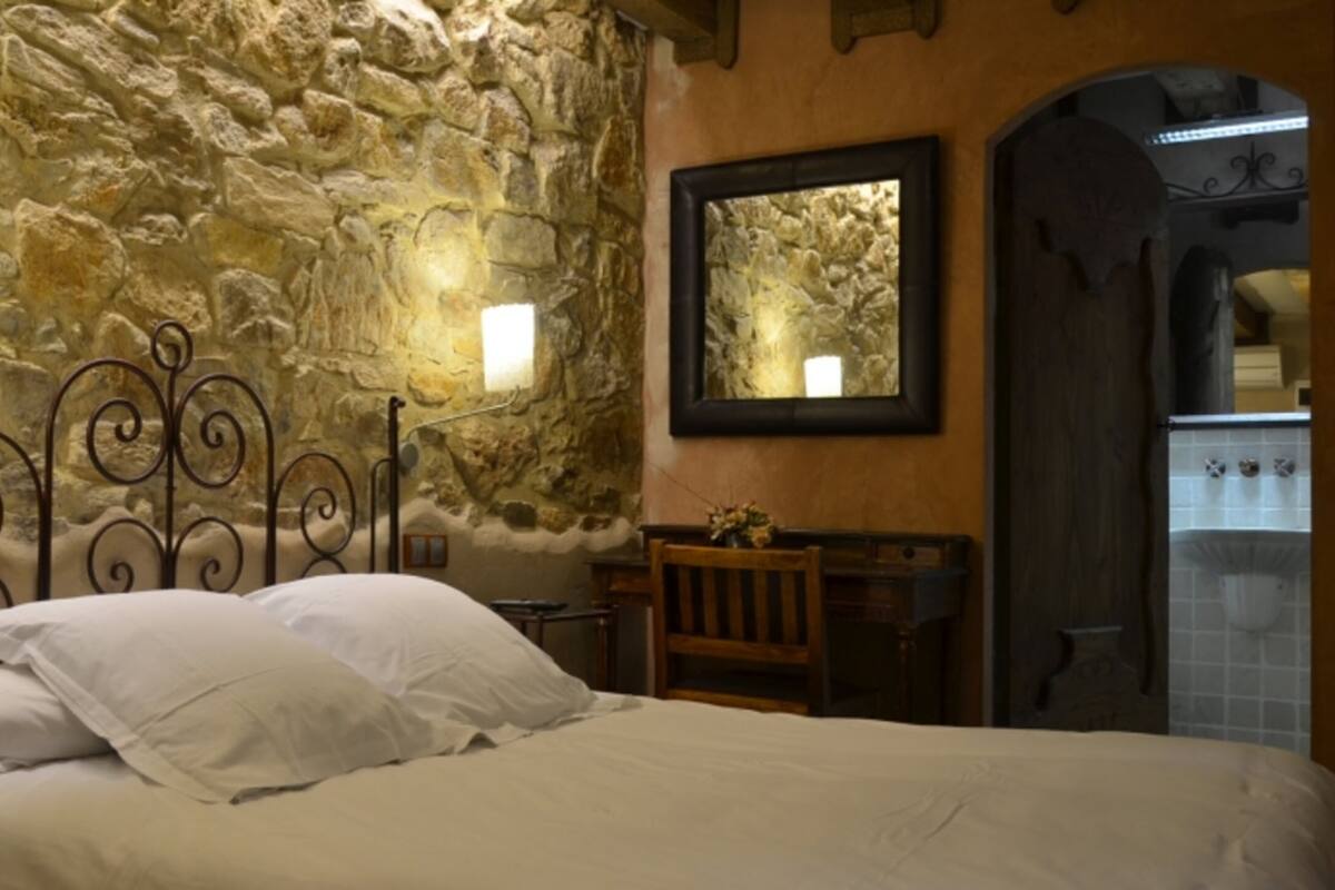 Ein Schlafzimmer mit Steinwand, einem Bett mit weißen Leinen, einem Holzstuhl, einem kleinen Tisch mit Lampe und einem Spiegel an der Wand.