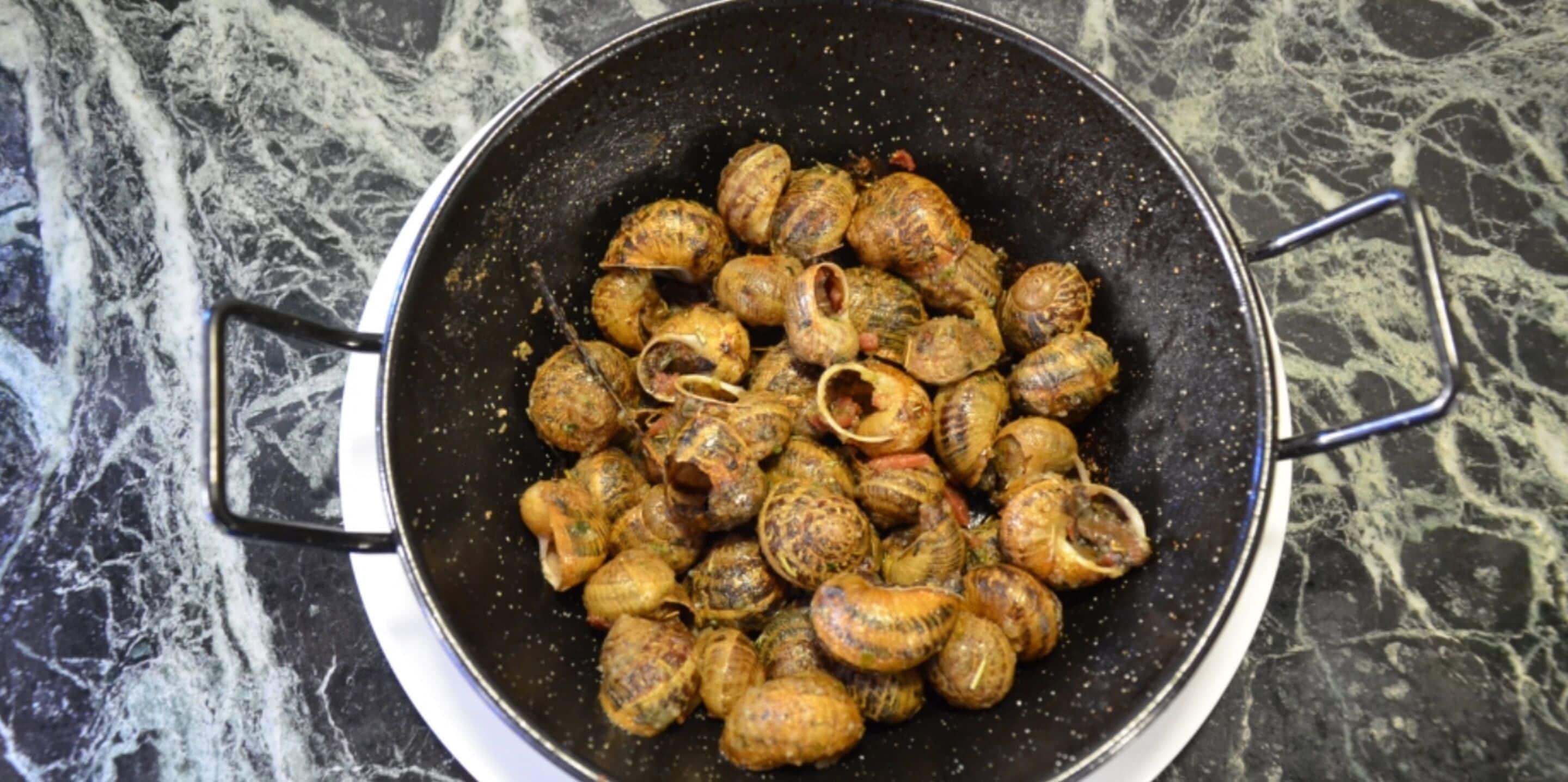 Een grote zwarte pan met gekookte slakken op een marmeren aanrecht.