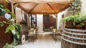 Um alpendre coberto com uma mesa e cadeiras de madeira, cercado por vasos de plantas e um barril rústico.