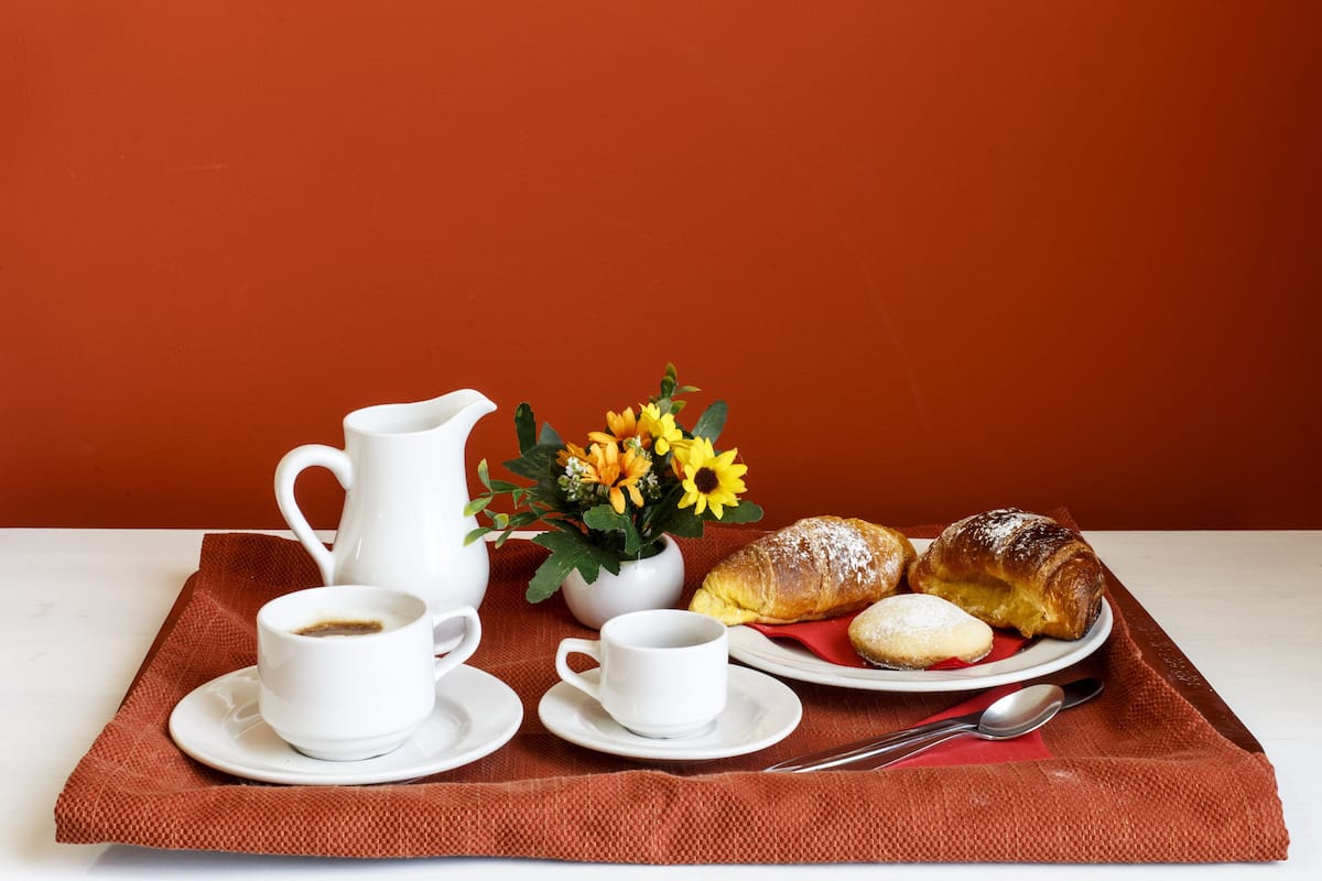 Une table dressée pour le petit-déjeuner avec une carafe, des tasses, un vase de fleurs et des viennoiseries sur une nappe.