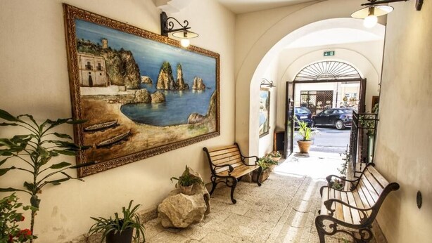 Um corredor com um quadro de uma paisagem costeira, bancos, plantas em vasos e um arco que dá para a rua.