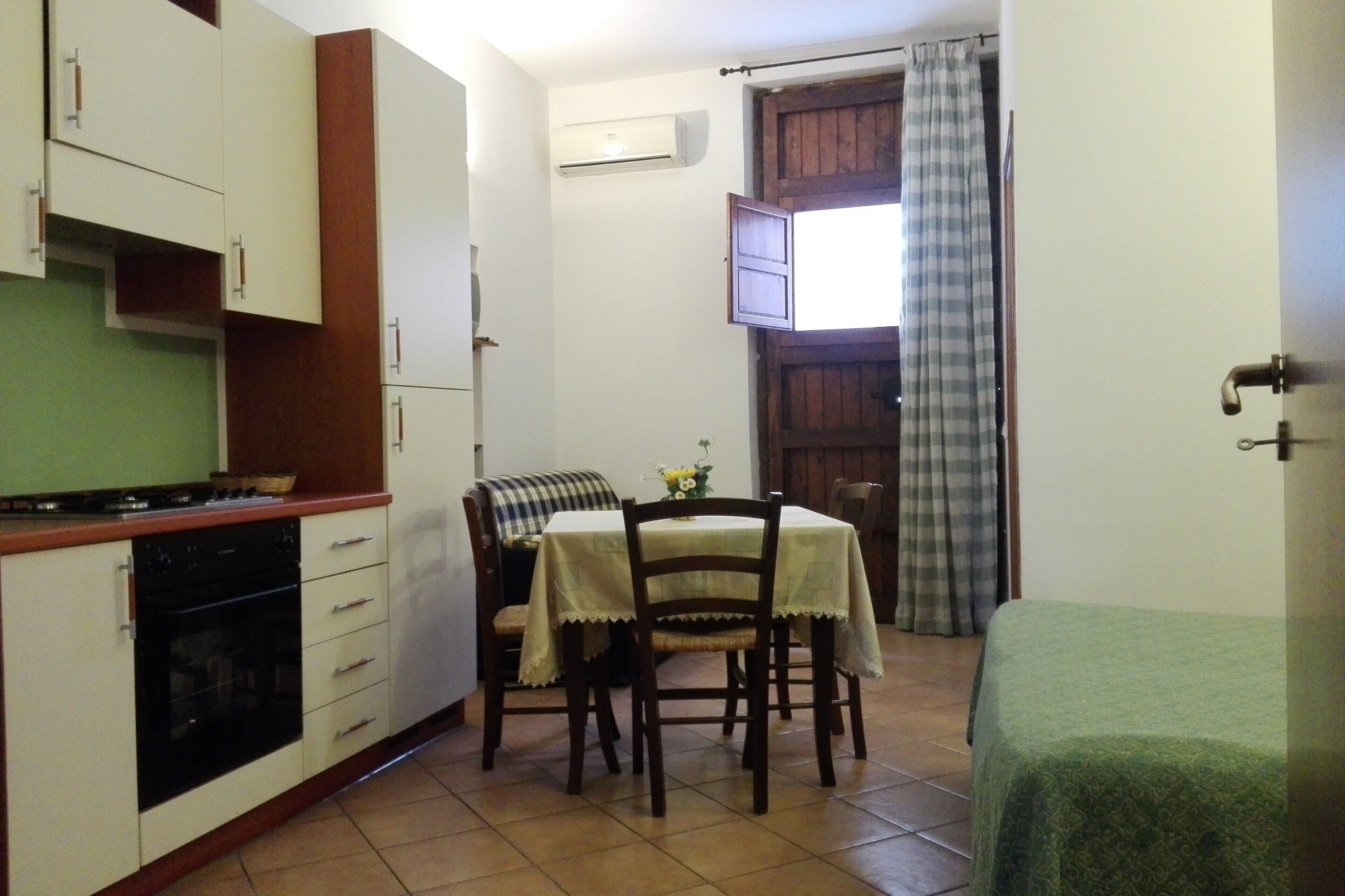 A compact kitchen with a stove, oven, and white cabinets. There is a dining table with chairs and a small table with a vase. A green bed is visible in the foreground.