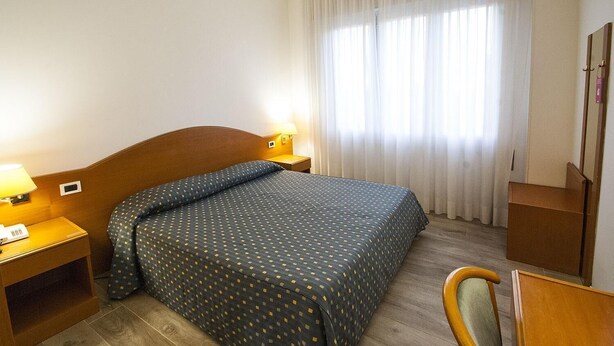 A hotel room with a bed, a nightstand with a lamp, a wooden chair, and a desk with a mirror.