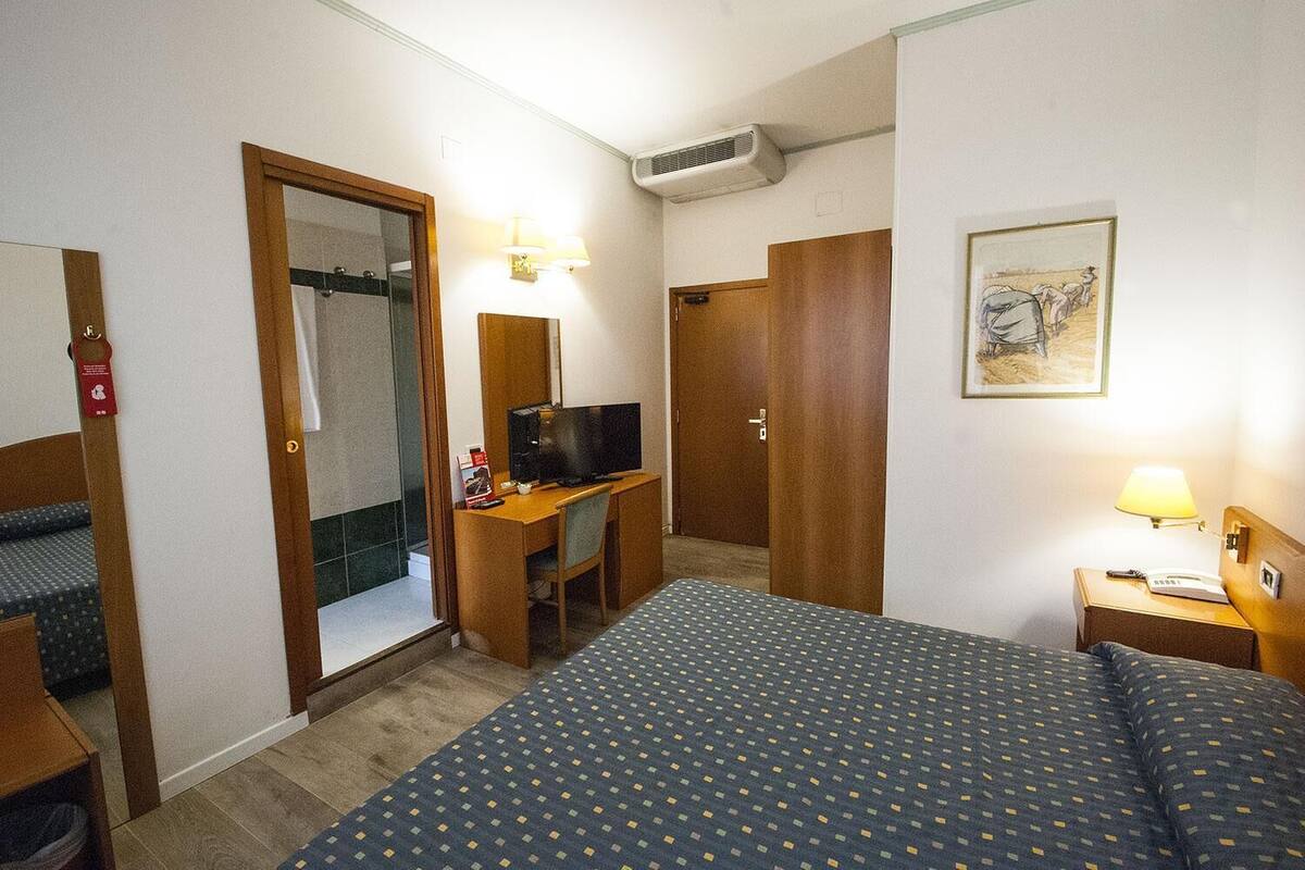 A hotel room with a bed, a desk with a TV, a bathroom with a mirror, and a painting on the wall.