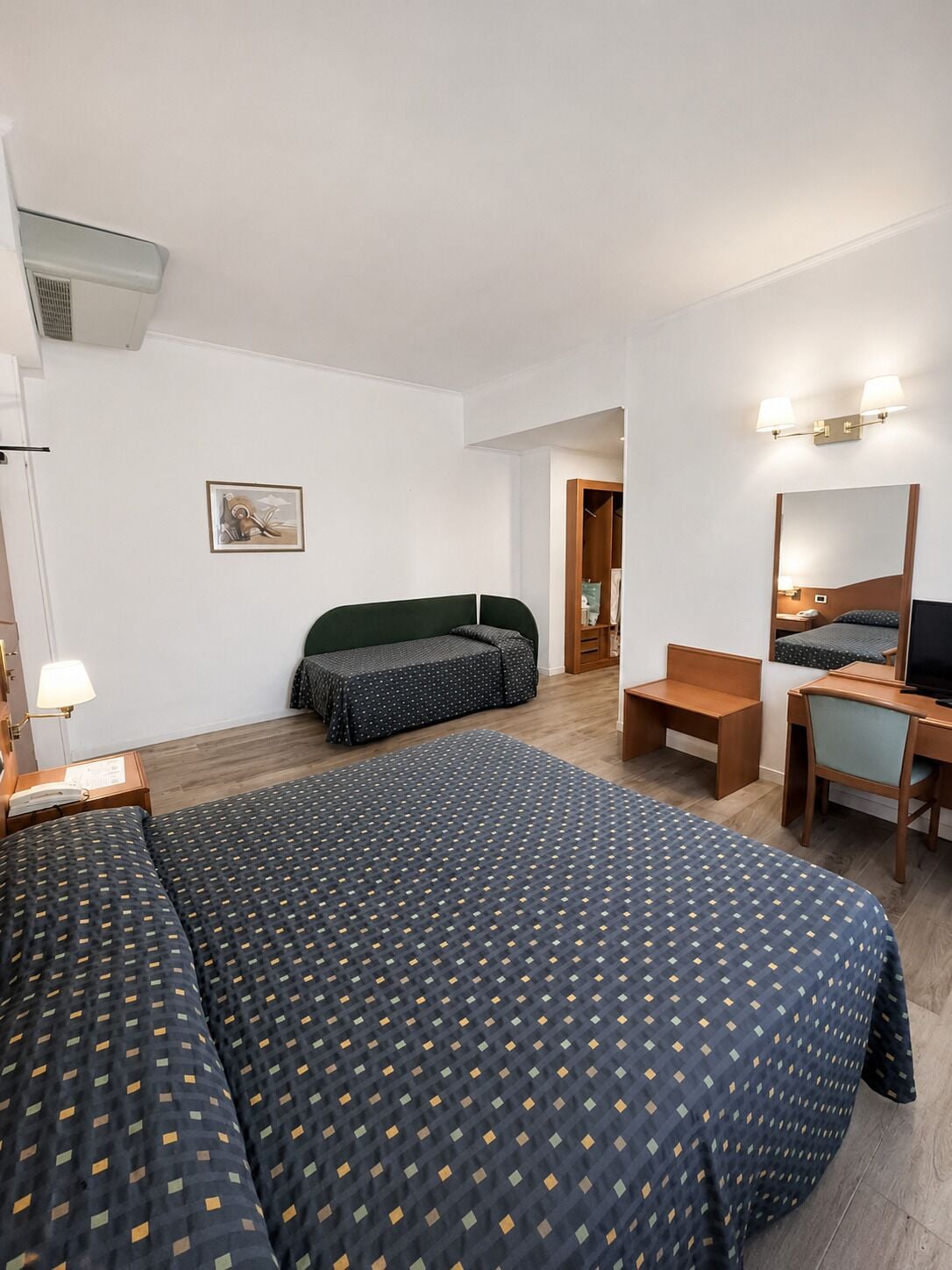 A hotel room with two beds, a desk, a chair, a mirror, a lamp, and a wardrobe.