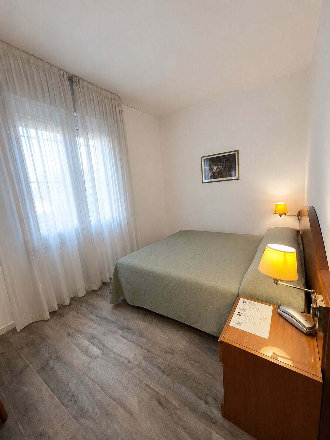 Habitación de hotel con cama, mesita de noche, lámpara, un cuadro en la pared y ventana con cortinas.